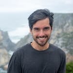 A smiling man in a black shirt poses in front of a scenic coastal cliff in Portugal.